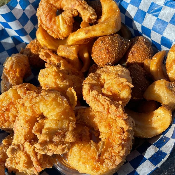 Fried shrimp, hush puppies, and potato wedges in a blue and white checkered paper basket.