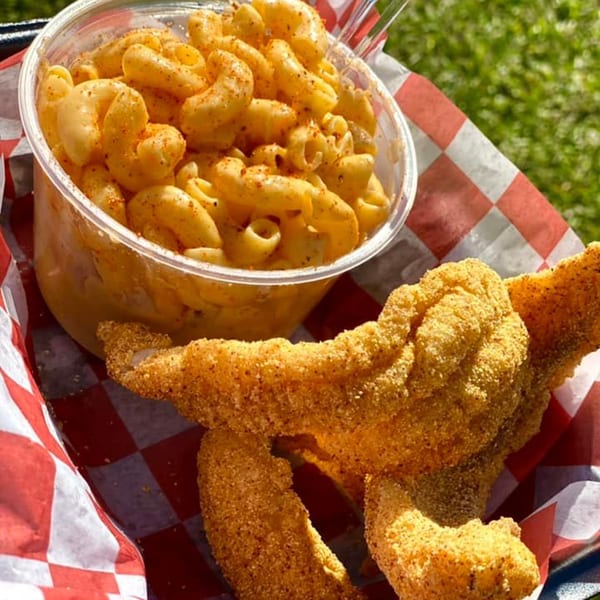 Fried catfish fillets and a clear cup of macaroni and cheese in a red and white checkered basket.